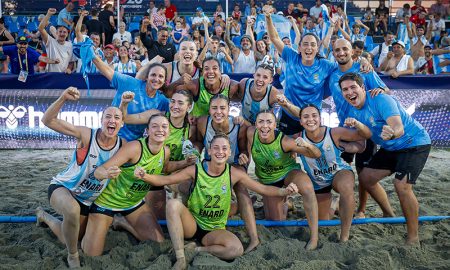 A beach handball team in blue and green uniforms celebrates on the sand with cheering fans in the background.