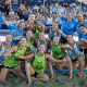 A beach handball team in blue and green uniforms celebrates on the sand with cheering fans in the background.