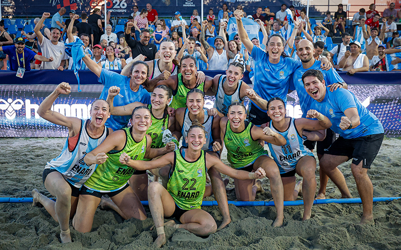 A beach handball team in blue and green uniforms celebrates on the sand with cheering fans in the background.